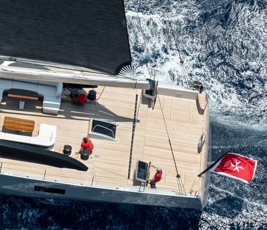 Aerial view of a sailing yacht deck with crew and a Maltese flag at the stern